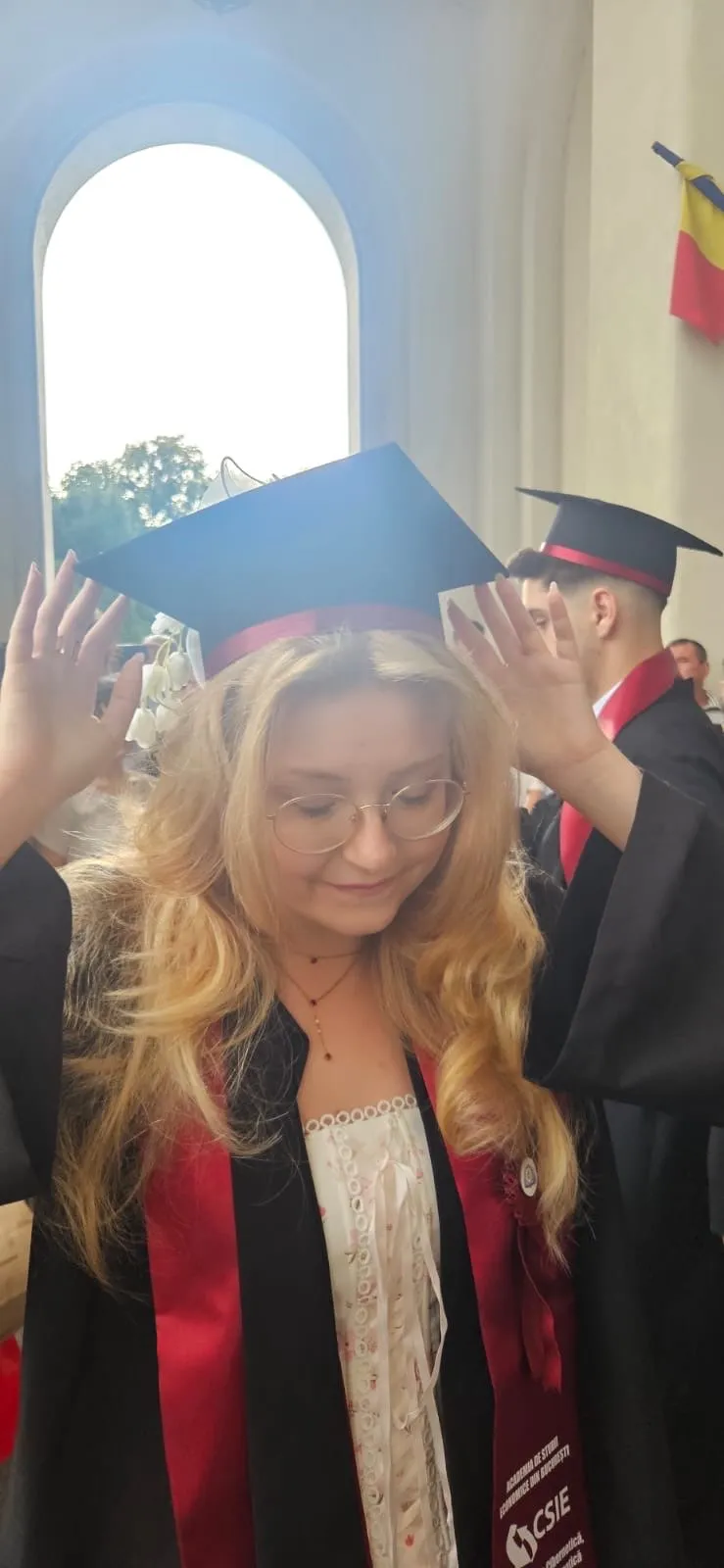 Alessia la absolvirea ASE București, în robă neagră și eșarfă roșie, aranjându-și tichia cu ambele mâini.