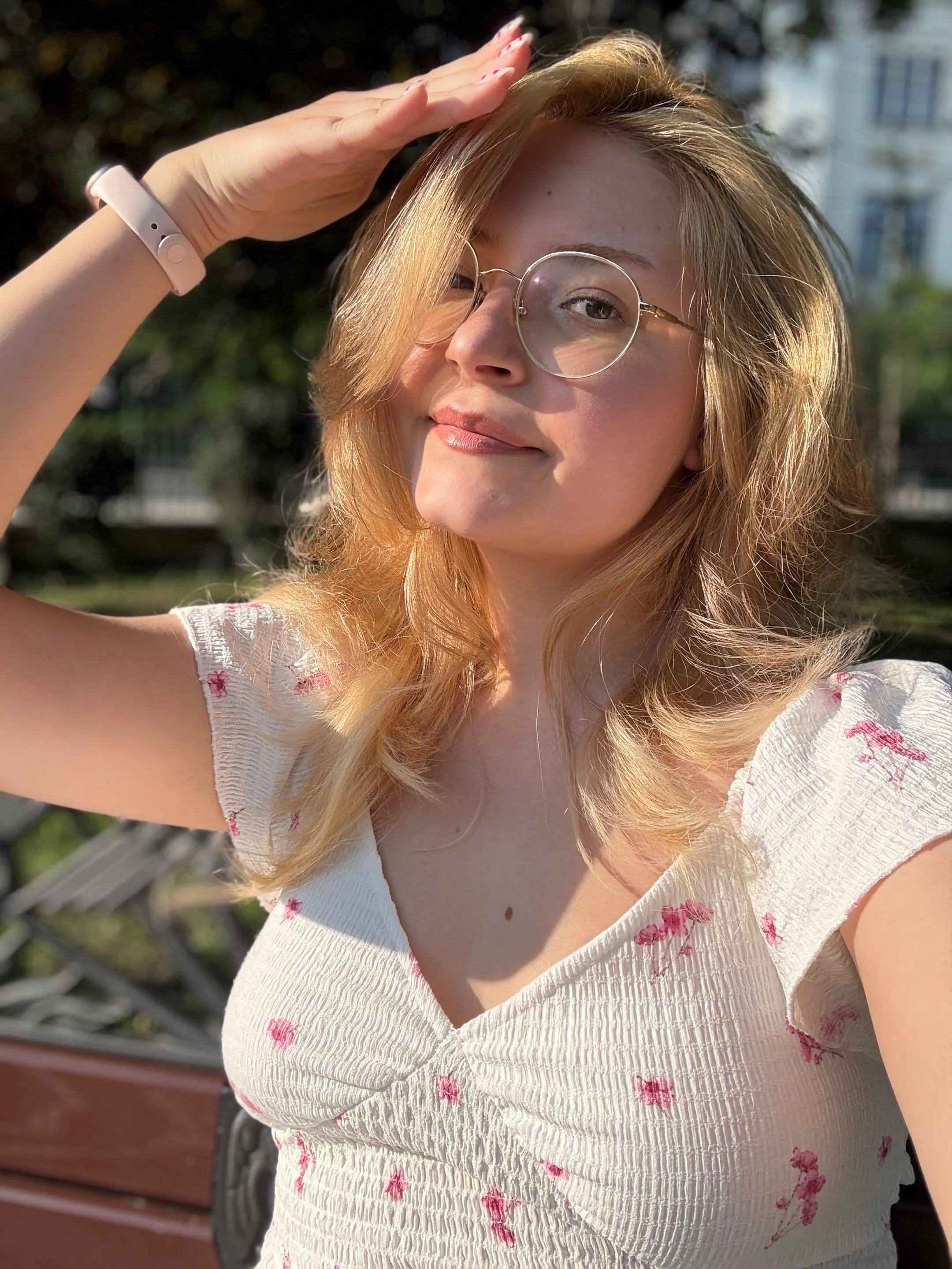 Alessia în soare, ferindu-și ochii cu o mână și zâmbind, cu ochelari rotunzi și o bluză albă cu flori roz mici.